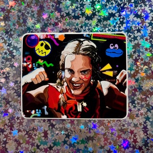 May include: A colorful sticker featuring a cartoon illustration of a woman with a determined expression. She is wearing a red and black outfit and has a yellow star on her cheek. The background is filled with geometric shapes and abstract designs.