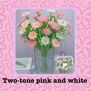 May include: A clear glass vase filled with two-tone pink and white daisy-like flowers. The flowers have yellow centers and green stems. The image also includes a small decorative box with a floral design.