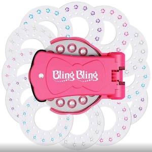 May include: A pink "Bling Bling" tool with a bow-shaped design, surrounded by white circular discs. The tool has a hinged mechanism and is adorned with rhinestones. The discs are decorated with colorful gems.