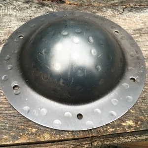 Può includere: Uno scudo rotondo in metallo nero con un centro a cupola e una superficie testurizzata. Lo scudo presenta una serie di piccole rientranze equidistanti attorno al bordo.