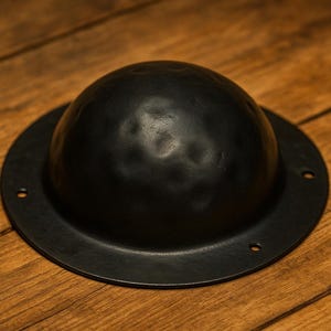 Op de afbeelding: Een zwart, koepelvormig metalen object met een cirkelvormige basis en drie gaten. Het object heeft een getextureerd oppervlak en staat tegen een houten achtergrond. Het logo "Lord of Medieval" is zichtbaar.