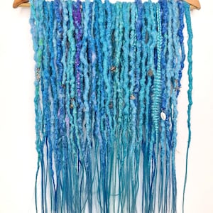 May include: A set of blue and teal synthetic dreadlocks with various embellishments, including beads and charms, hanging from a wooden hanger.