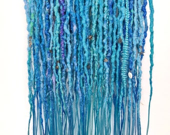 Op de afbeelding: Een set blauwe en turquoise synthetische dreadlocks met verschillende versieringen, waaronder kralen en bedels, hangend aan een houten hanger.