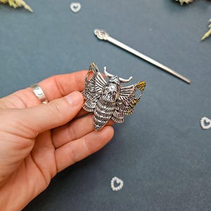 May include: A silver-toned metal hair accessory shaped like a moth with a skull design on its thorax. The moth features intricate wing detailing and a segmented body. A long, slender silver hair pin is positioned diagonally above the moth ornament.