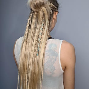 May include: A woman with long blonde hair styled in dreadlocks with blue and white accents. The hair is pulled back into a bun.