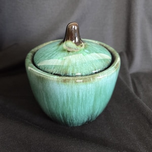 Puede incluir: Un azucarero de cerámica verde turquesa con tapa. El cuenco y la tapa tienen un acabado brillante. La tapa tiene un pequeño pomo marrón. El cuenco tiene forma redondeada.