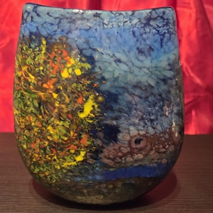 Könnte beinhalten: Eine dekorative Glasvase mit einem einzigartigen, farbenfrohen Design. Die Vase weist eine Mischung aus Blau, Gelb, Grün und Braun auf, die ein abstraktes, künstlerisches Muster erzeugt. Die Vase hat einen abgerundeten Boden und eine leicht gebogene Oberseite.