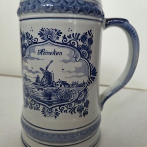 May include: A white ceramic beer stein with blue floral and windmill design. The stein has a handle and the word "Heineken" is displayed in a blue framed design. The stein is decorated with blue accents.
