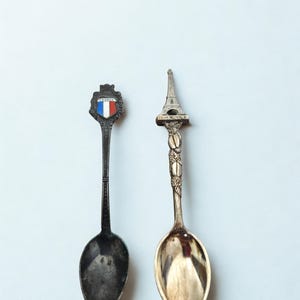 May include: Two vintage souvenir spoons. One spoon has a shield with the French flag colors and the word "FRANCE". The other spoon features a miniature Eiffel Tower. Both spoons have ornate handles and are made of metal.