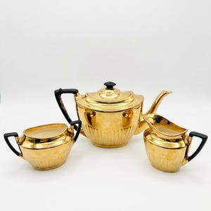 Könnte beinhalten: Goldfarbenes Teeservice mit Teekanne, Sahnekännchen und Zuckerdose. Die Teekanne hat einen schwarzen Griff und einen Deckelknopf. Sahnekännchen und Zuckerdose haben schwarze Griffe. Das Set hat ein geripptes Design.