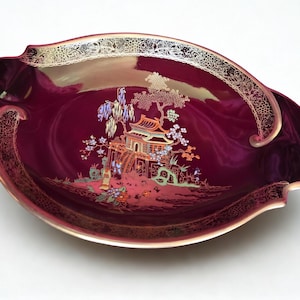 Könnte beinhalten: Burgunderfarbene dekorative Servierschale mit goldenem Rand und aufwendigen goldenen Details. Die Mitte zeigt eine detaillierte Illustration einer Pagode, Bäume und Figuren in einer Landschaftsszene. Die Schale hat eine einzigartige, geschwungene Form.