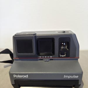 May include: A gray Polaroid Impulse instant camera with a black lens and flash. The camera has a red accent line and the word "Polaroid" and "Impulse" printed on the front. A black strap is attached to the side.
