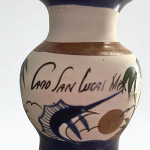 May include: A small, hand-painted ceramic vase with a flared rim and base. The vase features a blue, brown, and cream colour scheme. It has a stylized illustration of a fish and the text "Cabo San Lucas, MX".