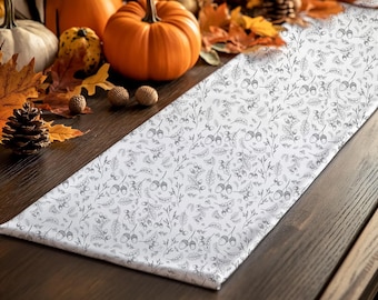 May include: White table runner featuring a grey pattern of acorns, leaves, and branches. The runner is displayed on a dark wooden surface, with pumpkins, autumn leaves, and pine cones in the background.