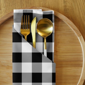 May include: A gold fork, knife, and spoon set on a black and white checkered napkin. The napkin is folded and placed on a wooden tray, creating a stylish table setting.