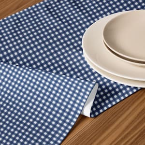 May include: A navy blue and white checkered table runner with a stack of three beige plates on a wooden surface. The runner has a classic gingham pattern, adding a touch of rustic charm to the table setting. The plates are stacked neatly, ready for a meal.