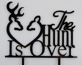 May include: Black acrylic cake topper with a heart shape formed by two deer heads and the text "The Hunt Is Over".