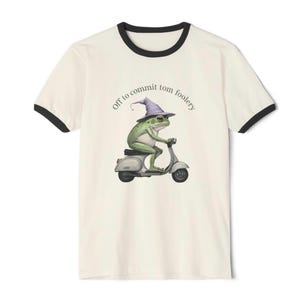 Peut inclure: T-shirt crème à bordures noires avec une illustration fantaisiste. Une grenouille verte portant un chapeau de sorcier violet conduit un scooter gris. Le texte "Off to commit tom foolery" est imprimé au-dessus de la grenouille.