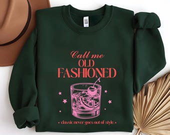 Peut inclure: Sweat-shirt vert foncé avec l'inscription "Call me OLD FASHIONED" en rose. Une illustration rose d'un cocktail Old Fashioned est en dessous, avec la phrase "classic never goes out of style."