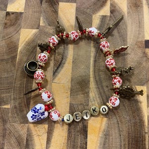May include: A beaded bracelet with white and red floral beads, gold-colored charms, and the name "JETKO" spelled out in gold letters. Includes a small teacup charm and a blue and white floral bead.