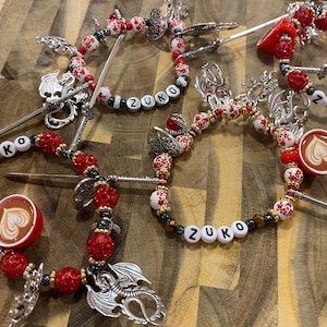 May include: Several beaded hair accessories with the name "Zuko" spelled out in white letter beads. The accessories feature red and white beads, silver charms, and small coffee cup charms with heart-shaped latte art.