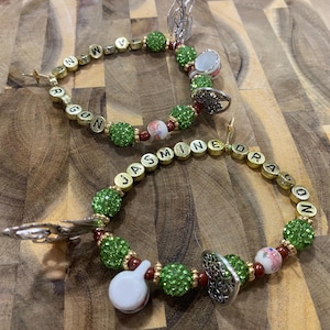 May include: Two beaded bracelets with the names "Jasmine Dragon" and "Dragon" spelled out in gold letter beads. The bracelets feature green, red, and white beads, along with silver charms and gold accents. The bracelets are on a wooden surface.