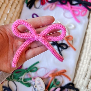 May include: A bright pink knitted bow hair clip with a textured yarn finish. The bow features two loops and two trailing ends, attached to a metal clip mechanism. Multiple other knitted bows in various colors are visible in the background.