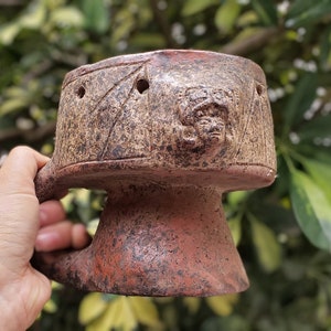 May include: A hand-held, rustic-style ceramic mug with a handle. The mug is primarily brown with reddish-brown accents and features a carved face and geometric patterns. The mug has several small holes near the top and is held against a blurred green background.
