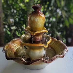 Puede incluir: Una fuente de cerámica de tres niveles con un esmalte marrón y dorado. La fuente tiene una cascada de agua y está diseñada para ser utilizada en interiores.