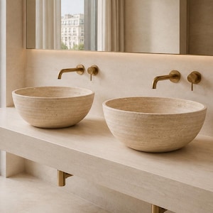 Puede incluir: Un tocador doble con dos lavabos de piedra beige, circulares y de sobreponer, colocados sobre una encimera de piedra a juego. Dos grifos de pared de latón cepillado con manijas a juego se sitúan sobre cada lavabo, junto a un gran espejo.