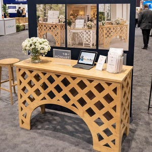 Puede incluir: Un stand de exhibición para Homart Architectural Wood & Iron Design. El stand presenta una mesa de madera clara con un diseño de celosía, una computadora portátil y un jarrón con flores blancas. El telón de fondo incluye el logotipo de Homart e imágenes de sus productos.