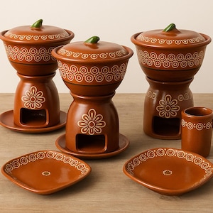 Puede incluir: Un juego de fondue de color terracota con tres ollas, tapas y soportes, además de una taza y dos platos cuadrados. Cada pieza presenta un diseño floral blanco y un patrón decorativo alrededor del borde. Las tapas tienen detalles verdes.