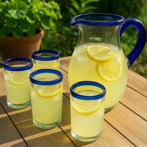 May include: A glass pitcher and four glasses filled with lemonade and lemon slices. The pitcher and glasses have a clear glass body with a blue rim. The drinks are on a wooden table, with greenery in the background.
