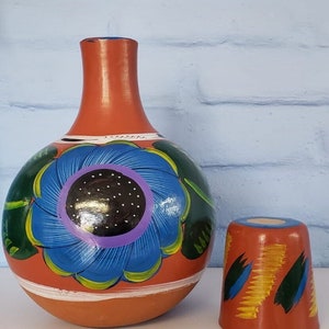 May include: Hand-painted terracotta bottle and cup set with a blue flower design. The bottle has a wide base and a narrow neck. The cup is smaller and has a similar design.