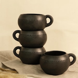 Puede incluir: Cuatro tazas de cerámica gris oscuro apiladas, con una quinta al lado. Cada taza tiene una forma redondeada, un asa pequeña y una línea de pequeños agujeros. Las tazas se exhiben sobre un paño de tono neutro.