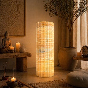 Puede incluir: Una lámpara de pie cilíndrica con un diseño texturizado y en capas. La lámpara emite un brillo cálido y amarillo, iluminando el espacio circundante. La lámpara es un elemento decorativo en una habitación con una paleta de colores neutros.