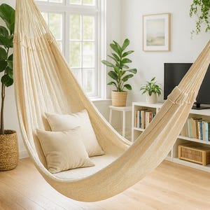 May include: Cream-colored hammock with two matching pillows, hanging indoors. The hammock is made of woven fabric and is suspended from a rope. The room has hardwood floors, a window, and several potted plants, creating a relaxing atmosphere.