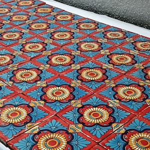 Puede incluir: Colección de baldosas cuadradas pintadas a mano con un patrón floral repetitivo. Cada baldosa presenta una flor central amarilla y roja rodeada de elementos decorativos azules y rojos. Las baldosas están dispuestas en una cuadrícula, creando una superficie vibrante y colorida.