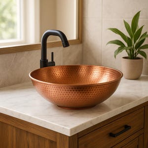 Könnte beinhalten: Ein kupferfarbenes Waschbecken mit Hammerschlag-Finish steht auf einer weißen Marmorplatte. Ein schwarzer Wasserhahn ist hinter dem Waschbecken montiert. Das Waschbecken steht auf einem Holzwaschtisch mit schwarzem Schubladengriff. Rechts steht eine Topfpflanze.