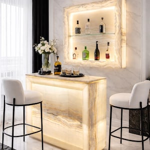 Puede incluir: Un bar de ónix retroiluminado con una estantería iluminada a juego que exhibe botellas de licor. Dos taburetes blancos con patas de metal negro están colocados frente a la barra. Un jarrón con flores blancas se encuentra sobre la barra, en una habitación con paredes y suelo de mármol.