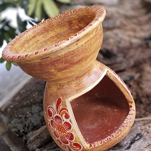 May include: A terracotta incense burner with a floral design in shades of red and orange. The burner has a wide, shallow top and a smaller opening on the side for burning incense.