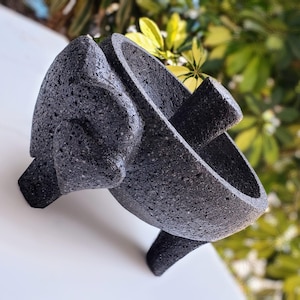 Puede incluir: Un molcajete de piedra volcánica negra con mortero y mano. El molcajete tiene tres patas y está hecho de piedra rugosa y texturizada.