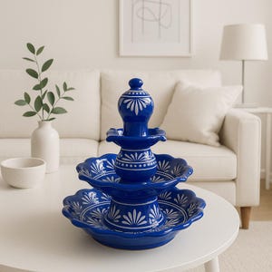 May include: A three-tiered, cobalt blue ceramic serving dish with white floral designs. The dish has a scalloped edge and a decorative top. The piece is displayed on a white table in a well-lit room.