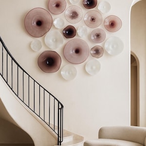 May include: A collection of decorative glass plates in varying sizes and shades of pink, white, and brown are mounted on a cream-colored wall. The plates have a unique, organic shape. A black wrought iron railing and a curved staircase are visible.