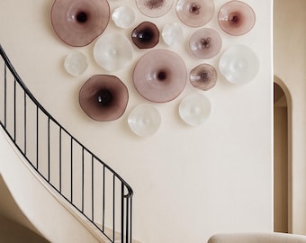 May include: A collection of decorative glass plates in varying sizes and shades of pink, white, and brown are mounted on a cream-colored wall. The plates have a unique, organic shape. A black wrought iron railing and a curved staircase are visible.