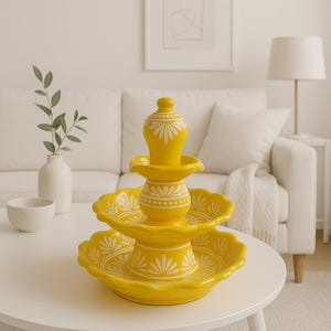 May include: A three-tiered yellow ceramic fountain with white floral designs. The fountain has a scalloped edge on each tier and a decorative finial. The fountain is a decorative home accent piece.