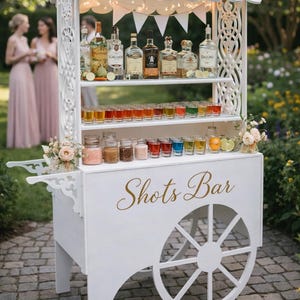 May include: A white wooden shots bar cart with a decorative roof, shelves, and a wheel. The cart is stocked with various bottles of liquor, shot glasses, and garnishes. The words "Shots Bar" are written in gold script.