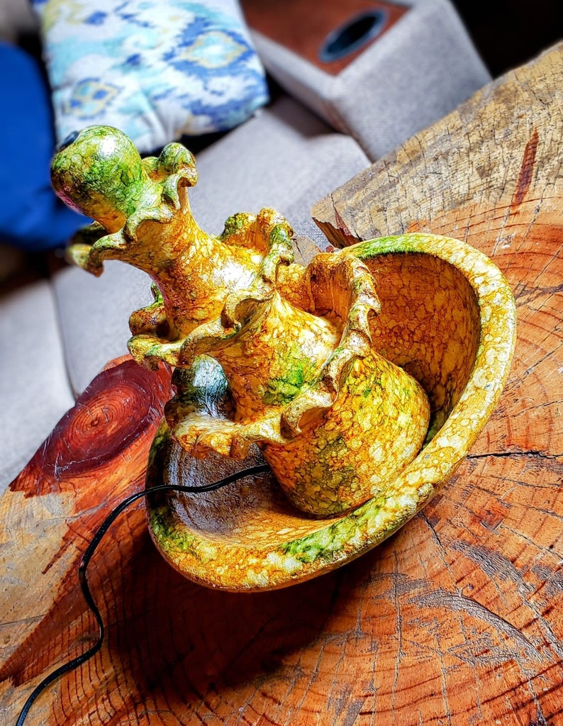 May include: A ceramic fountain with a multi-tiered design. The fountain is painted in shades of yellow, green, and brown, with a marbled effect. The fountain is sitting on a wooden surface.