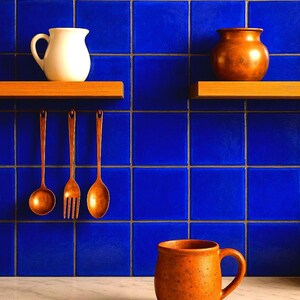 Peut inclure: Un pichet en céramique blanche et un pichet en cuivre sont posés sur des étagères en bois contre un mur carrelé bleu. Trois ustensiles en cuivre sont suspendus à des crochets au mur. Une tasse en céramique brune est posée sur un plan de travail en marbre blanc.