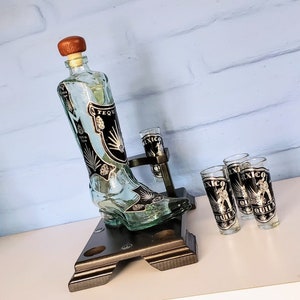 May include: A clear glass bottle shaped like a cowboy boot with a brown cork stopper. The bottle is decorated with black and white designs and the words "Tequila" and "Mexico". The bottle is sitting on a black wooden stand with four shot glasses. The shot glasses are clear glass and have black and white designs with the words "Mexico" and "Tequila".
